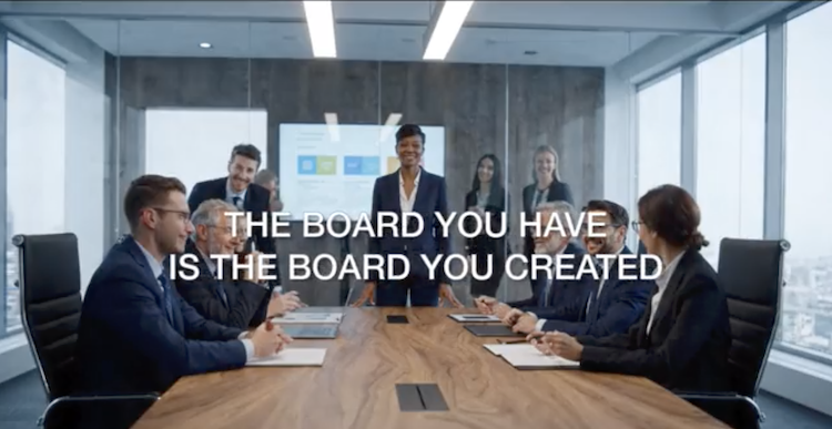 A wide-angle, medium shot of a diverse and dynamic corporate boardroom meeting in a modern high-rise office, featuring nine business professionals. The center figure is a smiling African American woman leader standing behind a long wooden conference table, with other diverse executives seated around it and several others, including men and women, standing behind her, creating a supportive group composition. Large windows provide a view of a city skyline. Overlaid in large, white, centered text is the phrase: "THE BOARD YOU HAVE IS THE BOARD YOU CREATED".